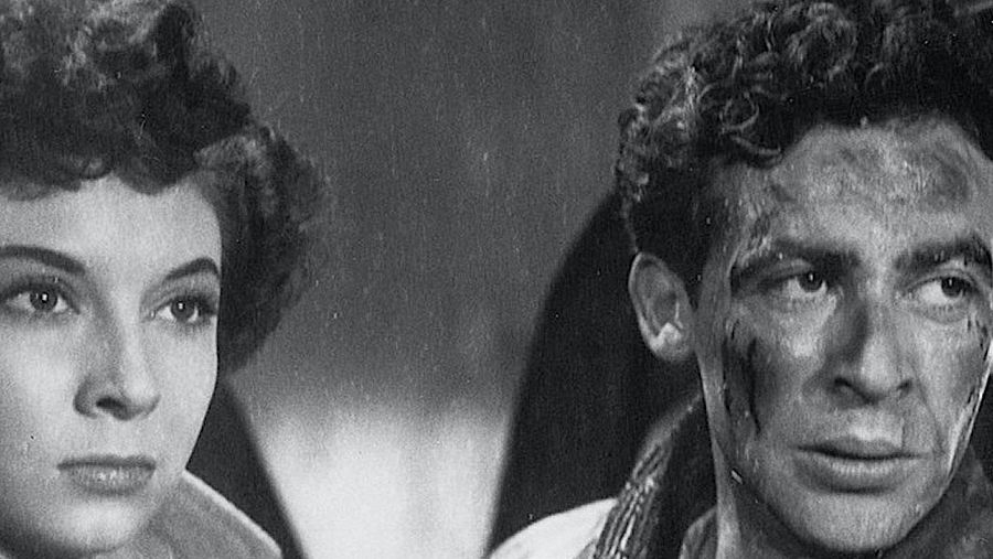 Imagen en blanco y negro: primer plano de dos rostros serios, un hombre con cabello rizado y manchas en la cara, y una mujer con cabello corto. Posible escena de película.