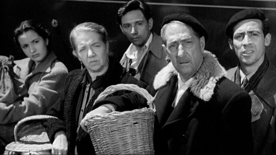 Grupo de seis personas, con cestas, en una estación. Expresiones serias, vestimenta sencilla.  Imagen en blanco y negro que transmite pobreza.