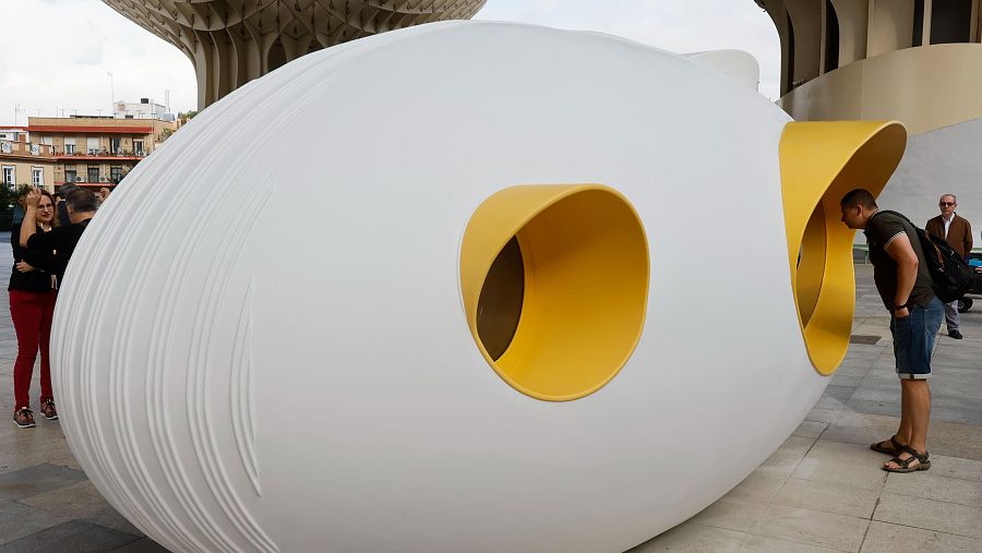 Escultura blanca, alargada y curva, con dos aberturas circulares amarillas. Personas la observan y fotografían en una plaza.