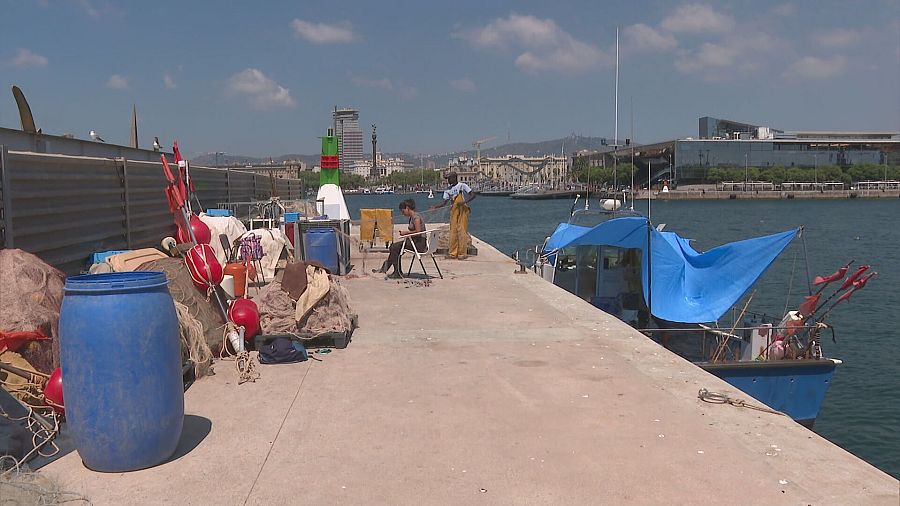 Escena en el Puerto de Barcelona: mujer arreglando redes de pesca cerca de un barco azul y aparejos.  Ambiente tranquilo.