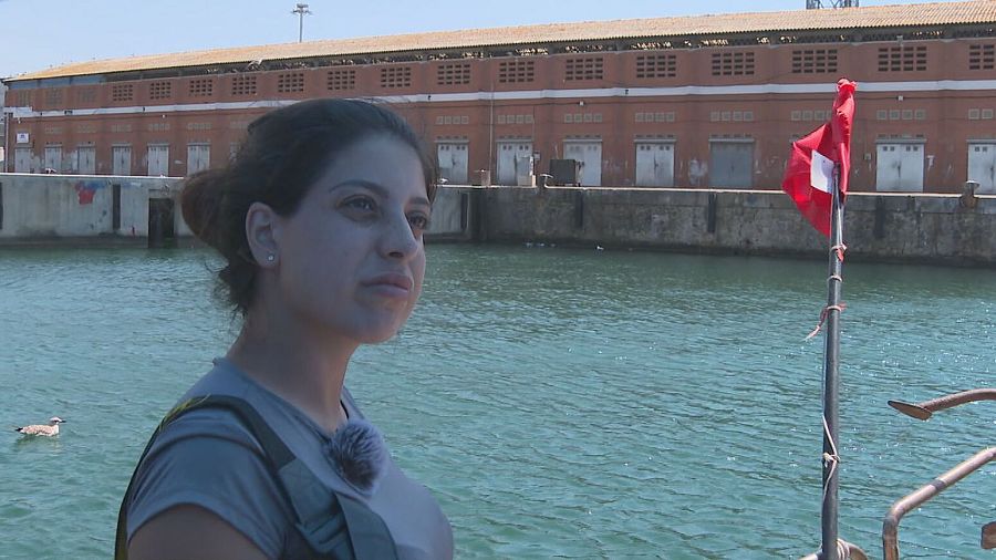 Mujer con micrófono en un muelle, mirando al mar con expresión seria.  Al fondo, un edificio y una bandera. Atmósfera tranquila.