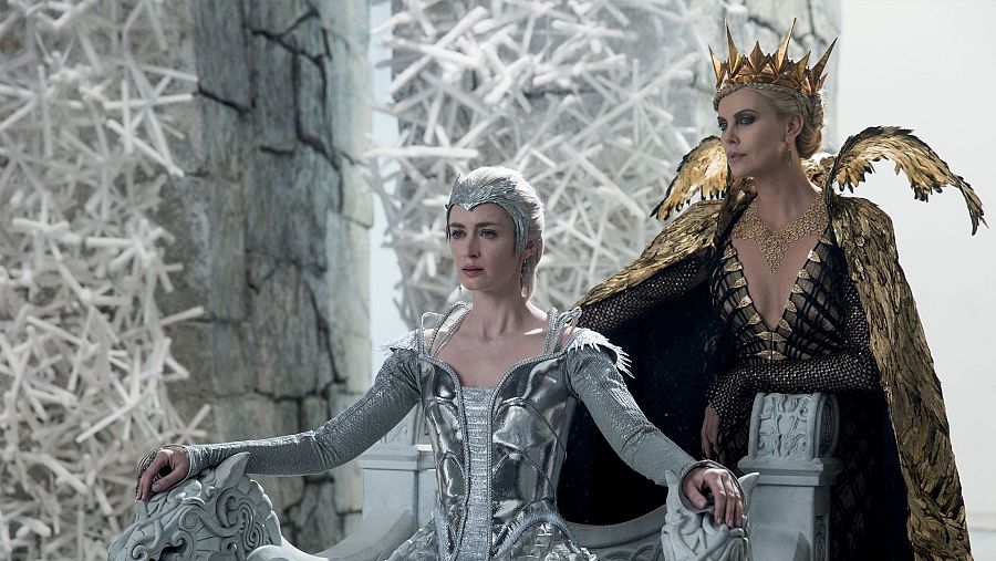 Escena de película con Emily Blunt en vestido plateado y Charlize Theron con atuendo negro y dorado y corona, sentadas en tronos en un suntuoso escenario de palacio.