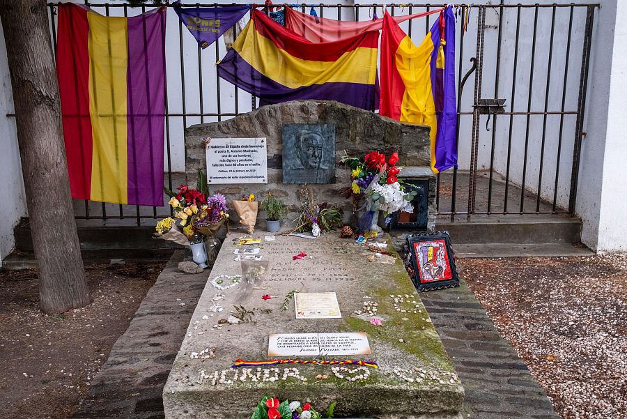 Tumba de piedra con placa de relieve, flores y objetos conmemorativos. Banderas republicanas españolas en la verja cercana. Atmósfera de respeto y recuerdo.