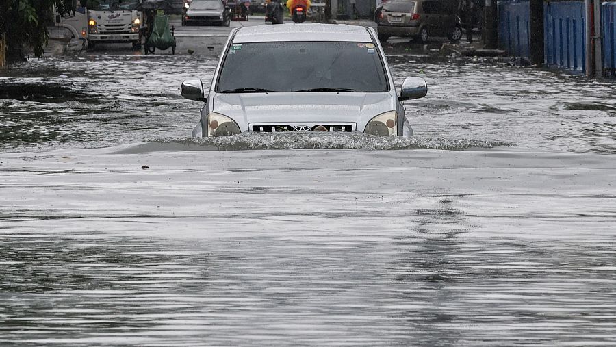 Coche SUV plateado semi-sumergido en agua turbia tras una inundación urbana.  Se observan otros vehículos y edificios al fondo.