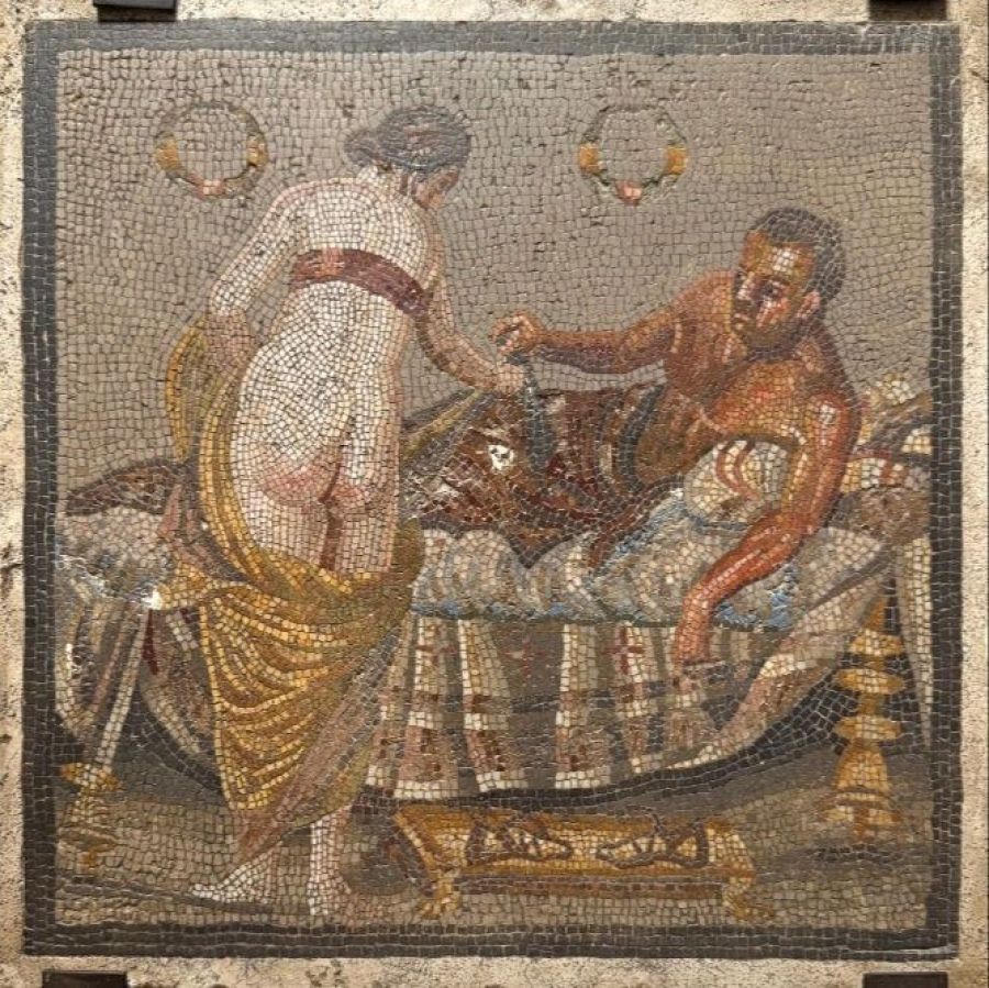 Mosaico romano erótico (siglo I a.C.) recuperado en Pompeya. Representa una escena íntima con figuras femeninas y masculinas parcialmente vestidas, junto a una cama con telas y coronas.
