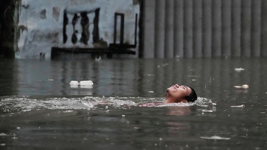 Hombre flotando en aguas de inundación, con desechos flotando a su alrededor. El agua llega casi al tope de una estructura cercana.