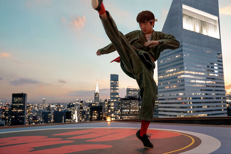 Joven en gi verde oliva realiza patada alta en azotea con plataforma circular; fondo urbano con edificios iluminados al atardecer.