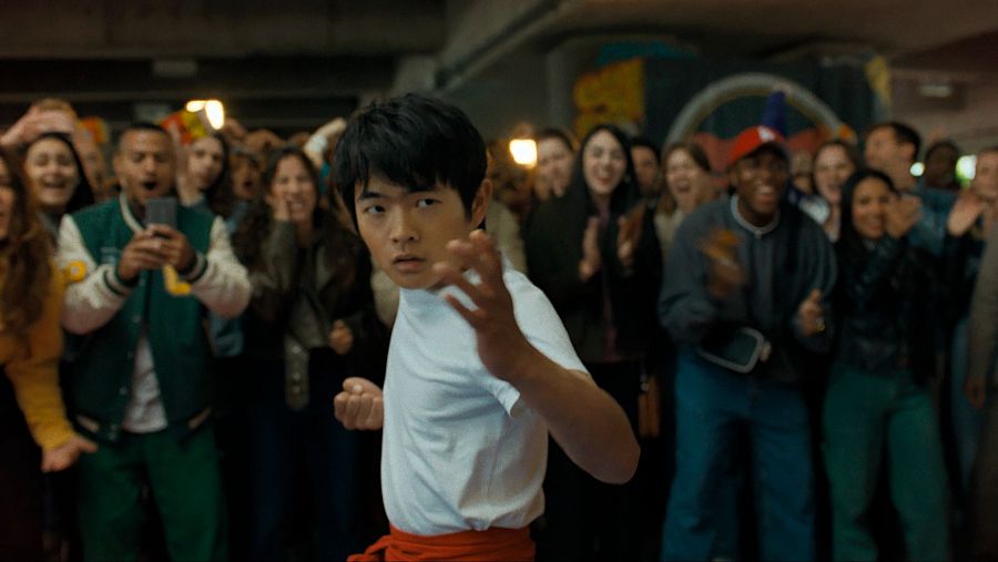 Joven en pose de karate, camiseta blanca y cinturón rojo, observado por un público diverso en un espacio interior con graffitis.