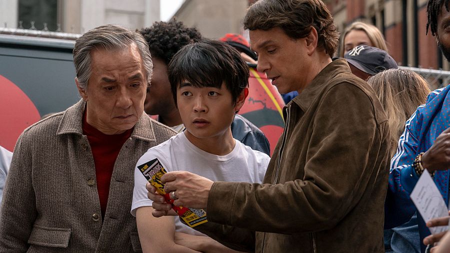 Escena de 'Karate Kid: Legends': tres personajes, uno muestra un ticket a otro, mientras un tercero observa. Ambiente urbano.