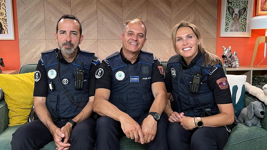Tres policías uniformados, con chalecos antibalas, sonríen en un sofá de un estudio de televisión de decoración moderna y colorida.