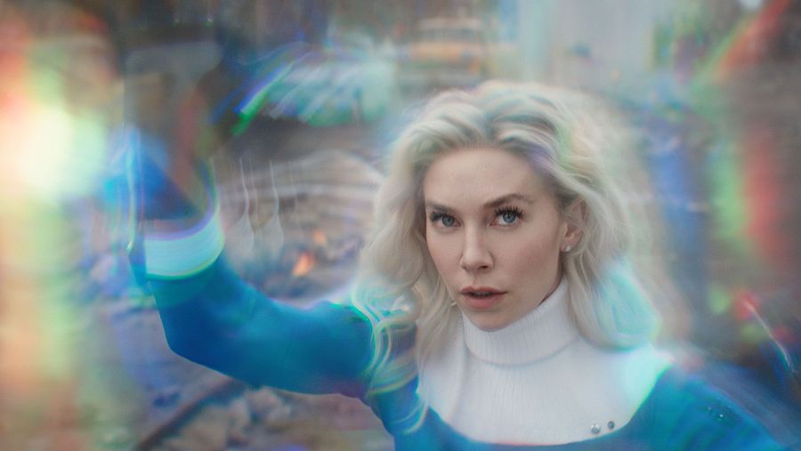 La Mujer Invisible, con traje azul y jersey blanco, proyecta un campo de fuerza iridiscente desde su mano en una escena de destrucción.