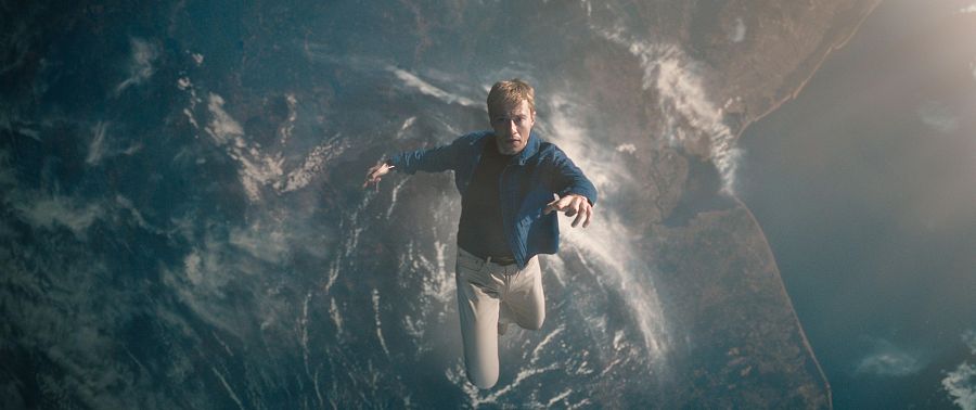 Hombre flotando en el espacio, con la Tierra visible al fondo. Viste camisa azul oscuro y pantalones beige; expresión seria, brazos extendidos.