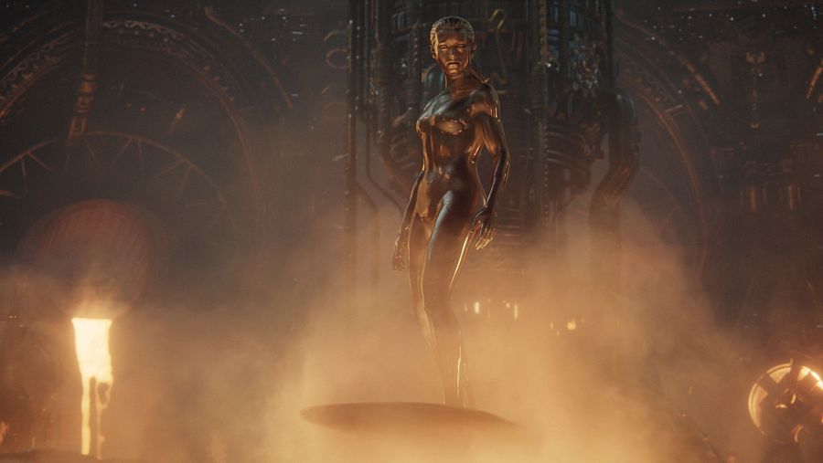 Figura femenina dorada, de pie sobre plataforma circular en ambiente oscuro e industrial, iluminada desde abajo por luces o llamas.  Expresión seria.