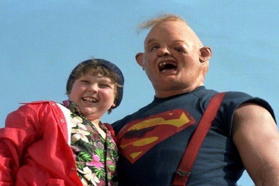 Chunk y Sloth, personajes de 'Los Goonies', sonríen en este fotograma. Chunk viste una chaqueta roja y Sloth una camiseta azul con tirantes.