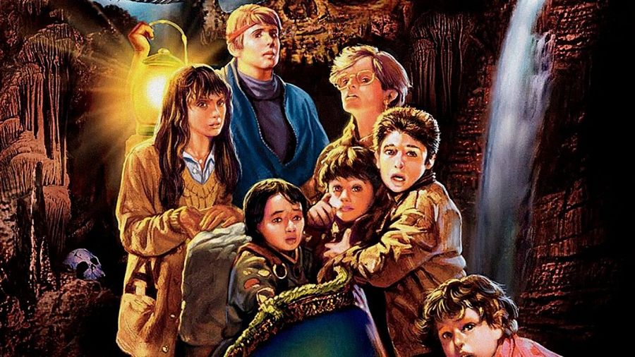 Niños exploran una cueva oscura con una cascada al fondo; uno se agarra el pecho, otro mira con aprensión. Una calavera y una lámpara iluminan la escena.