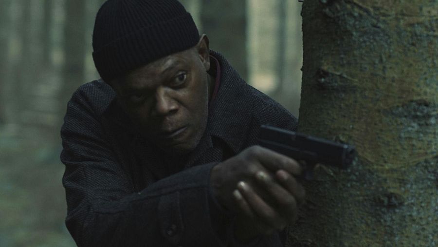 Actor agazapado tras un árbol en un bosque oscuro, empuñando una pistola.  Expresión seria y concentrada; atmósfera de suspense.