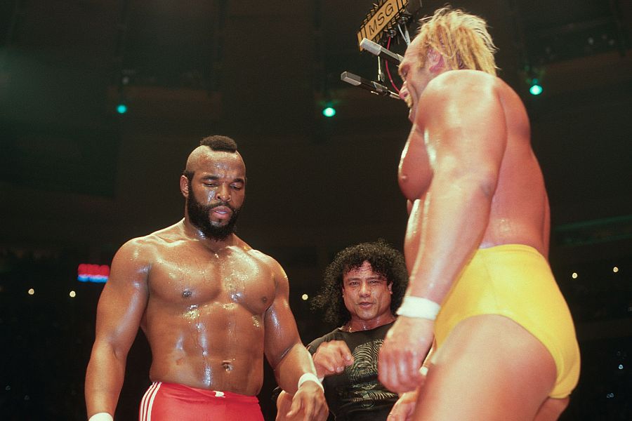 Intenso combate de lucha libre entre dos luchadores, uno con pantalones rojos y otro con bañador amarillo, en un gran recinto.