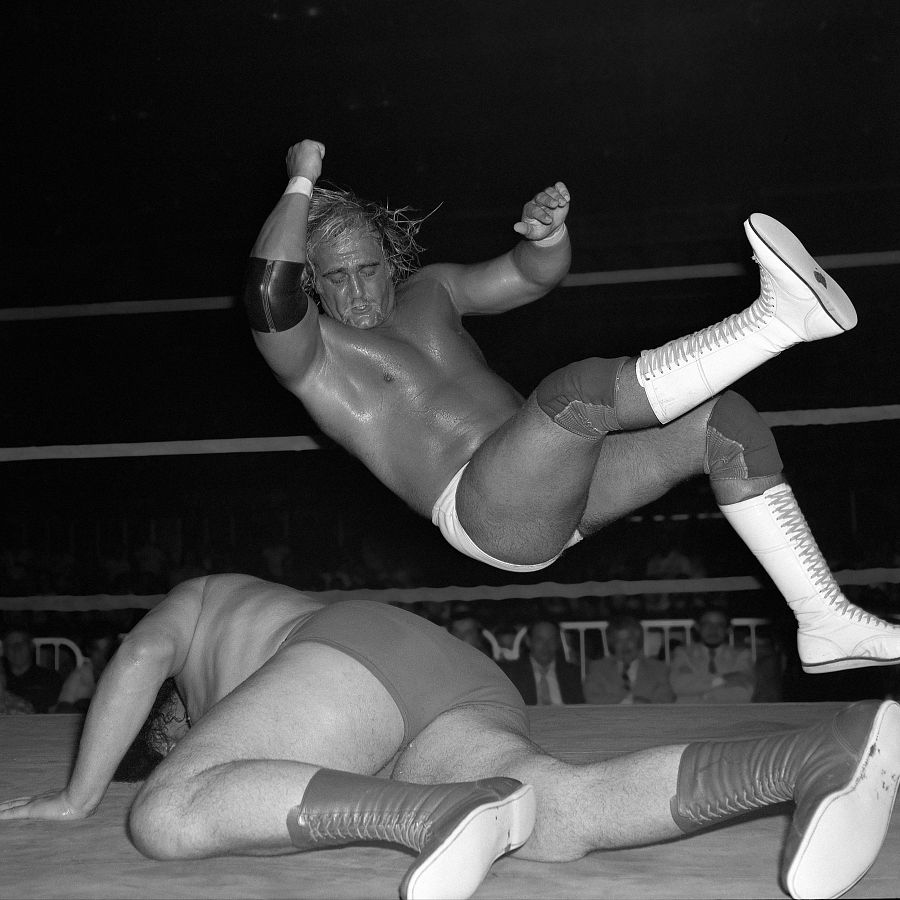 Imagen en blanco y negro de un combate de lucha libre entre dos hombres. Uno está en el aire, el otro en la lona. Público visible al fondo.