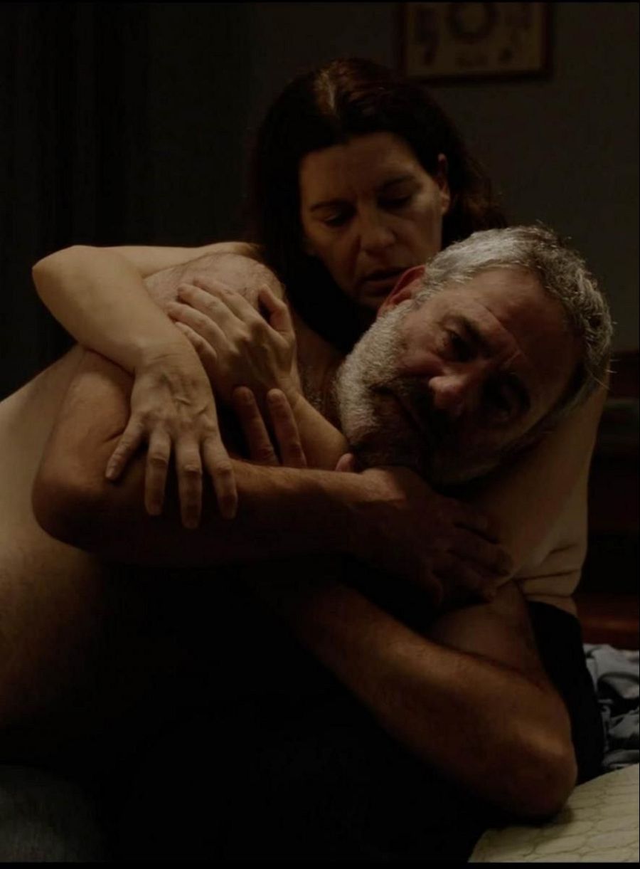 Pareja abrazada en cama, escena nocturna de una película. Iluminación tenue, ambiente íntimo y melancólico.  Detalles de ropa de cama y un cuadro en la pared.