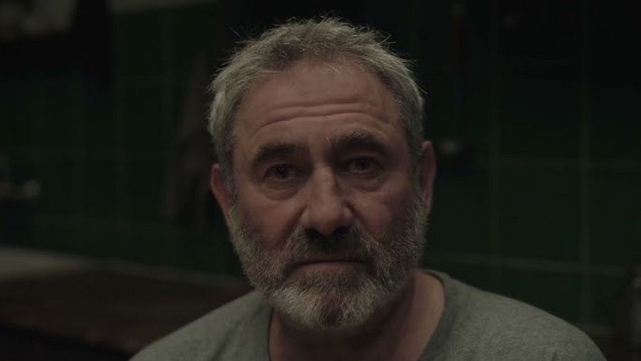 Raúl Arévalo, primer plano, pelo canoso, barba gris, expresión seria, camiseta gris, fondo con taquillas verdes oscuras, iluminación tenue.