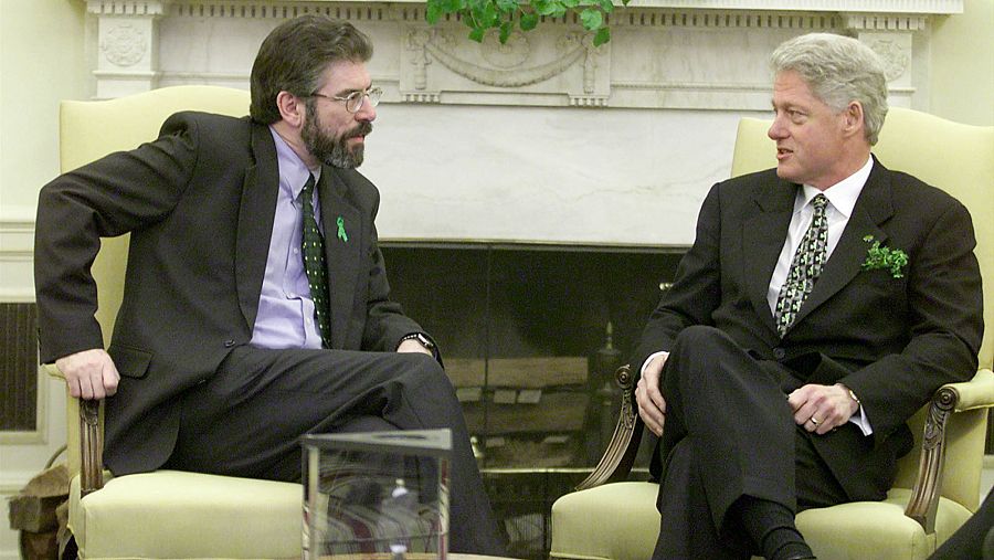 Reunión entre dos hombres en trajes oscuros, sentados en sillones de una sala formal con chimenea. Uno lleva una corbata con tréboles, el otro una corbata verde con lunares.