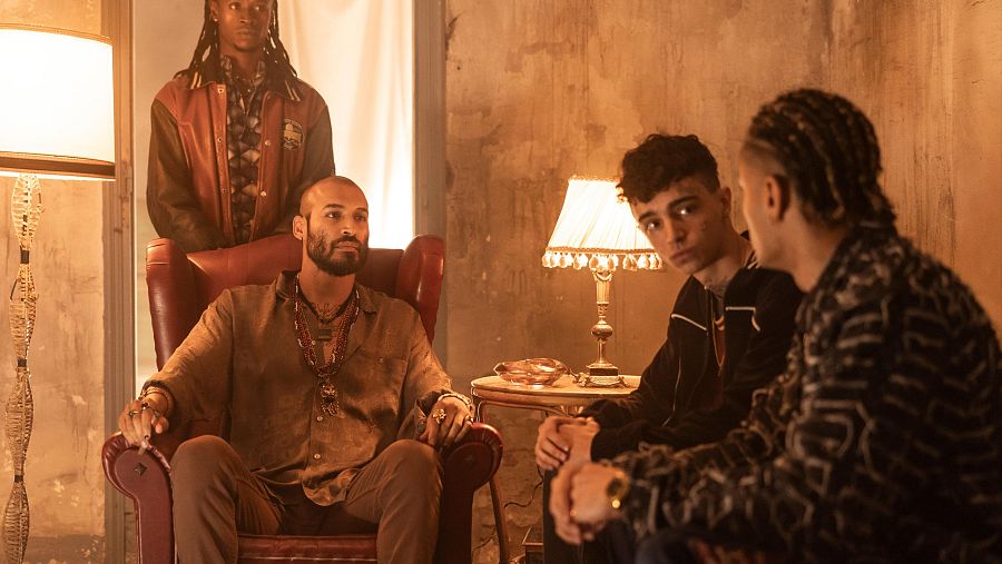 Escena oscura e íntima con cuatro hombres: uno calvo con barba en un sillón, dos jóvenes sentados frente a él y otro de pie al fondo. Iluminación cálida y ambiente misterioso.