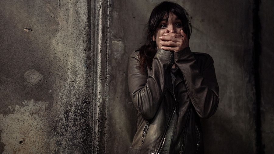 Mujer con expresión de terror, manos en la boca, chaqueta oscura y pelo oscuro hasta los hombros, frente a una pared de hormigón gris oscuro y con iluminación tenue.