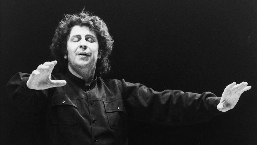 Mikis Theodorakis, en blanco y negro, dirige una orquesta con gesto concentrado; camisa oscura, cabello oscuro y rizado; fondo negro.