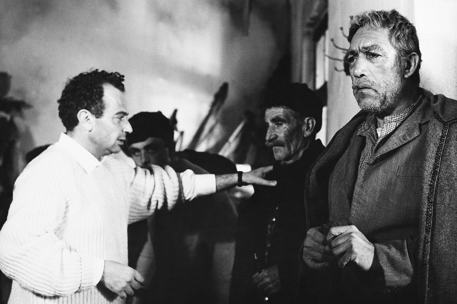 Escena de rodaje en blanco y negro: el director, con suéter claro, dirige a un actor con abrigo oscuro.  Ambiente rústico.