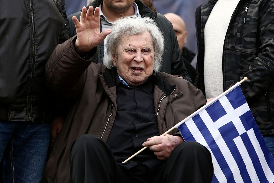 Mikis Theodorakis, en 2018, sentado y saludando con una bandera griega en la mano. Viste chaqueta marrón y camisa oscura.