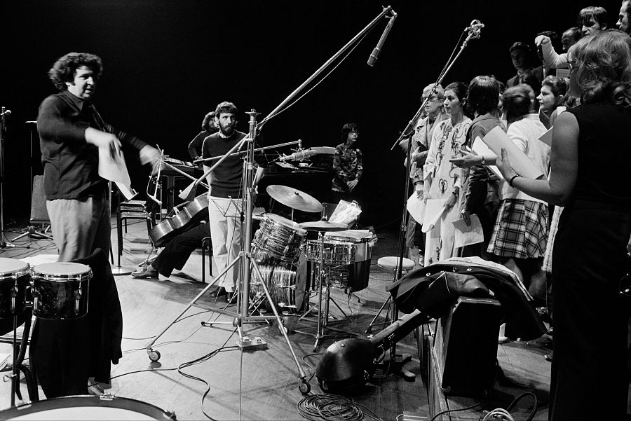 Mikis Theodorakis dirige una orquesta en París, 1970.  Imagen en blanco y negro mostrando al compositor gesticulando con una partitura, rodeado de músicos y un coro.