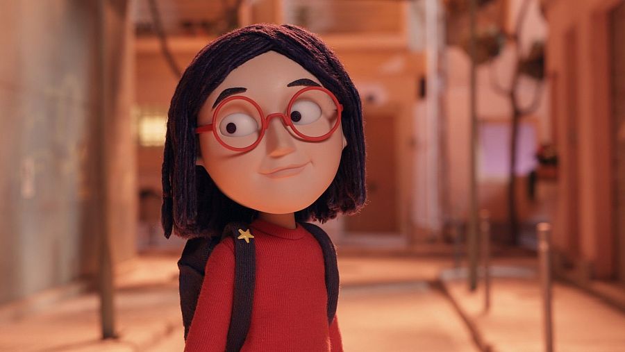 Niña con jersey rojo y mochila, gafas redondas rojas y pelo negro lacio hasta el cuello. Fondo desenfocado con tonos cálidos.