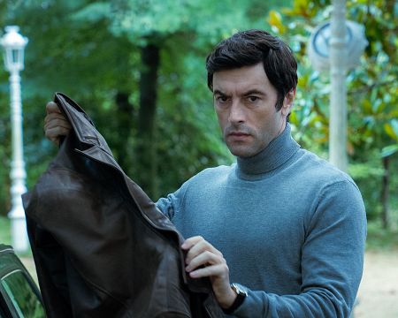 Hombre con jersey gris y chaqueta marrón en un parque, expresión seria, fondo con árboles y farola.  Ambiente tranquilo y misterioso.