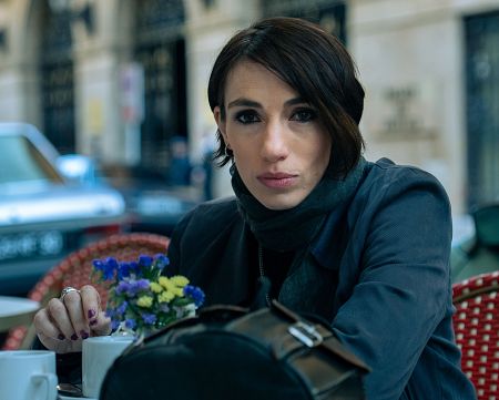 Actriz con chaqueta oscura y bufanda gris sentada en terraza de café, mirando a cámara con expresión seria.  Sobre la mesa hay tazas y flores.