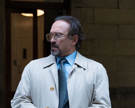 Hombre de mediana edad con gabardina beige y gafas, expresión seria, frente a una entrada. Texto superpuesto sugiere relación con comercio y frontera.