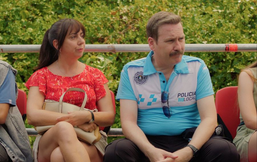 Dos adultos sentados en una grada observan un evento. Ella viste blusa roja; él, camiseta 