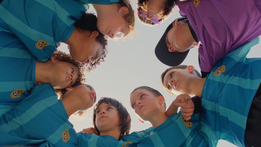 Niños y niñas con camisetas de fútbol, la mayoría azul turquesa con franjas, forman un círculo abrazados.  Imagen desde abajo, mostrando unidad.