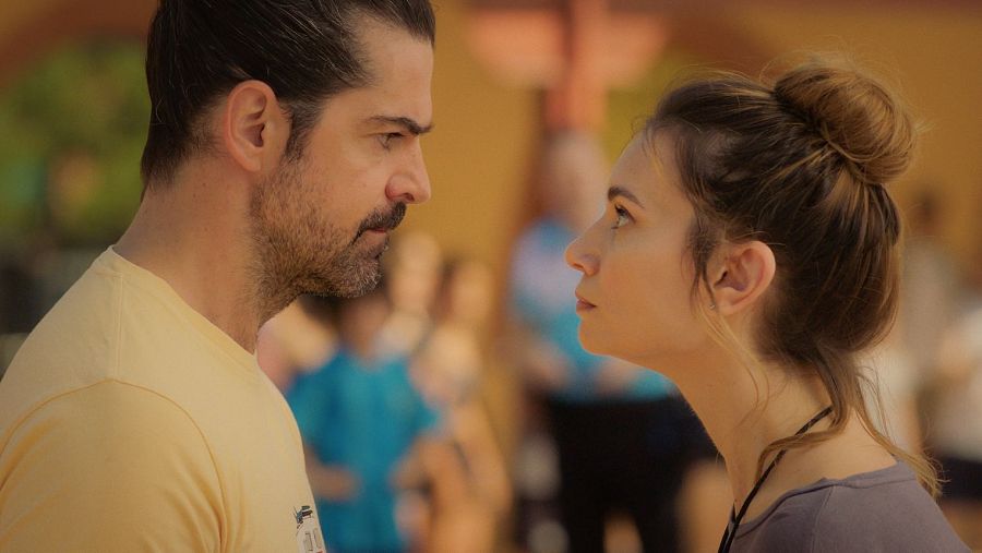 Un hombre y una mujer, con el pelo recogido en moños, se miran con seriedad en un patio o campo deportivo. Al fondo, figuras borrosas con ropa deportiva.