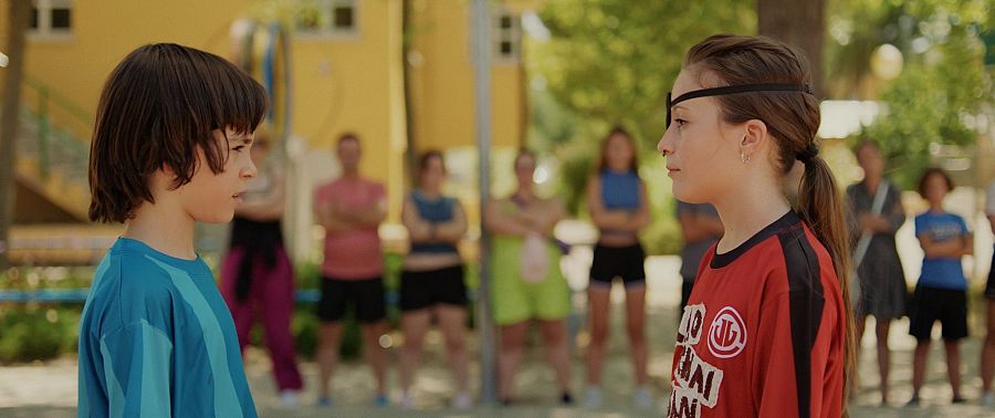 Dos niños, uno con camiseta azul-verde y otro con camiseta roja y antifaz, se enfrentan en un fotograma de 