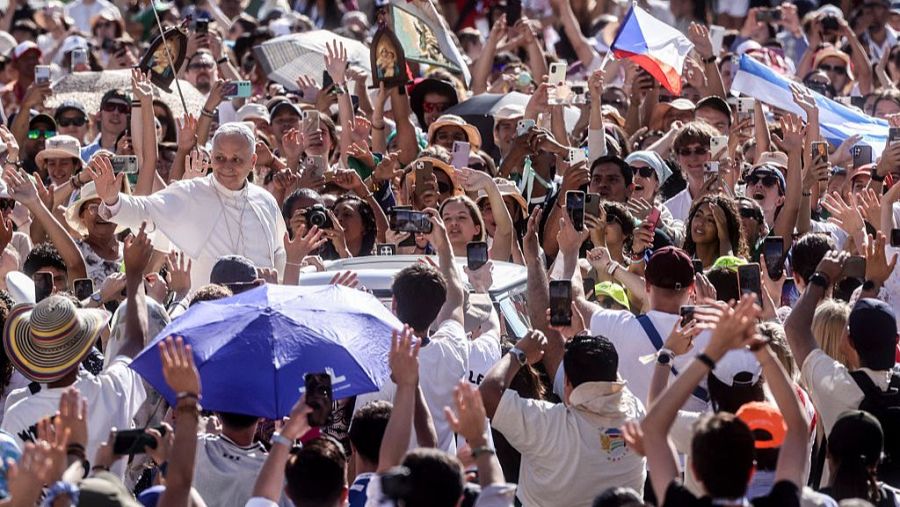 Multitud diversa rodea al Papa Francisco en un evento exterior.  Algunos llevan teléfonos y banderas; un paraguas azul destaca.