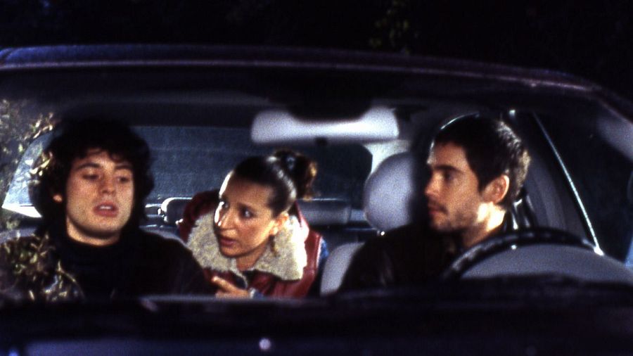 Interior de coche de noche. Tres personas conversan; el conductor mira al frente, uno escucha y otro habla.