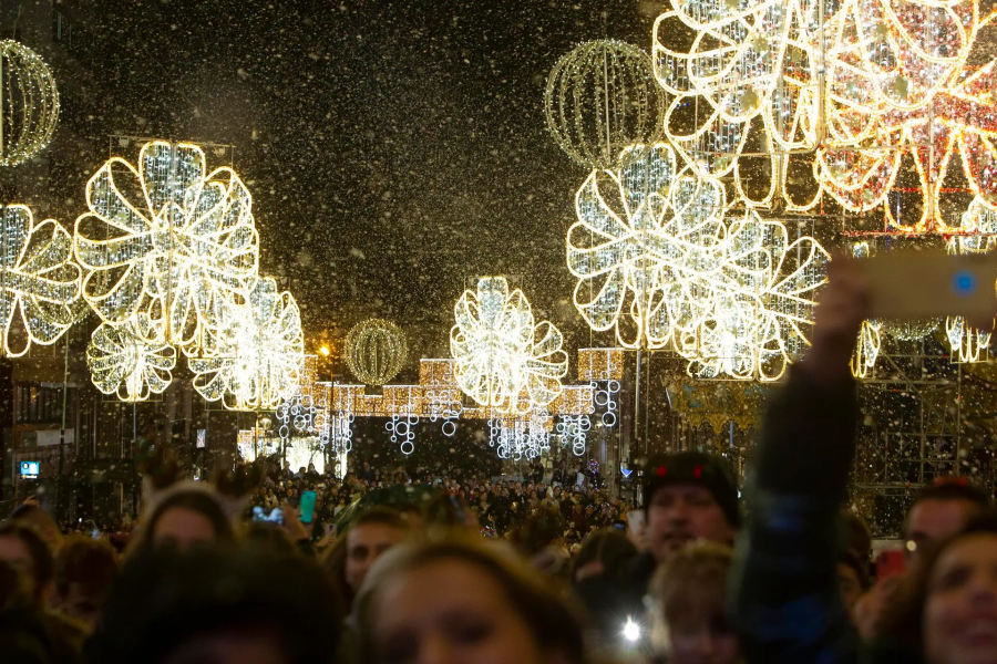 Iluminación navideña en una calle, con grandes estructuras de luz en forma de flores y esferas luminosas más pequeñas.  Multitud de personas, algunas grabando con sus móviles, bajo una nevada artificial.