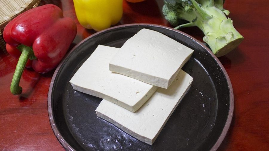 Plato con tres lonchas de tofu blanco, rectangulares y superpuestas, sobre superficie de madera.  Acompañado de pimientos y brócoli.