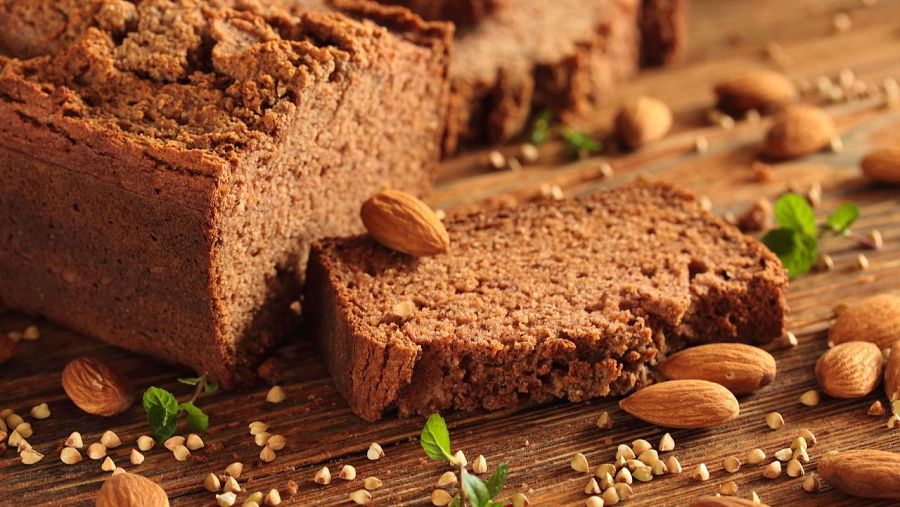 Pan integral sin gluten, húmedo y denso, con una rebanada cortada.  Alrededor hay almendras, granos y hojas verdes.