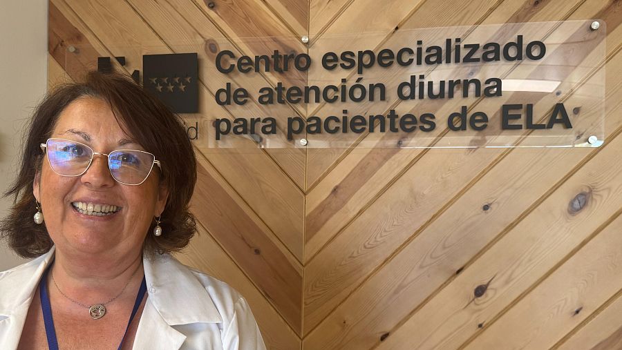 Mujer con bata blanca sonríe junto a una placa que anuncia un centro de atención diurna para pacientes de ELA.  Ambiente cálido con paredes de madera.