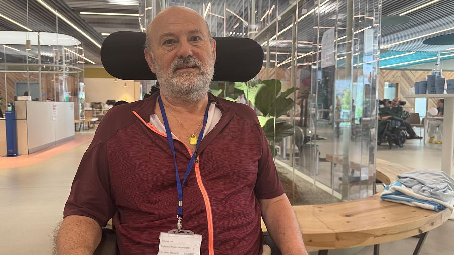 Hombre mayor en silla de ruedas, camiseta granate, en sala amplia con otros usuarios y paneles divisores. Prenda azul sobre mesa cercana.