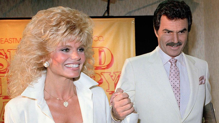 Loni Anderson y Burt Reynolds, sonrientes y tomados de la mano, en una foto de 1987. Ella luce camisa blanca y él traje blanco con corbata rosa; fondo con cartel amarillo y letras rojas.