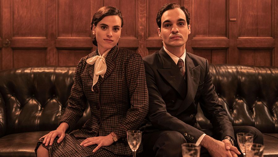 Escena de 'Karmele': hombre y mujer, en traje, sentados en un sofá, con copas en una mesa cercana.  Ambiente formal y clásico.