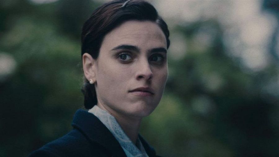 Imagen de Lola Dueñas con expresión seria, blusa clara y chaqueta oscura, pelo recogido y pequeños pendientes. Fondo desenfocado con vegetación.