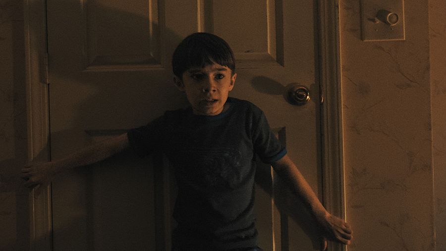 Niño con expresión de miedo agarrado a una puerta blanca en una habitación oscura. Viste camiseta oscura y tiene el pelo corto y oscuro.